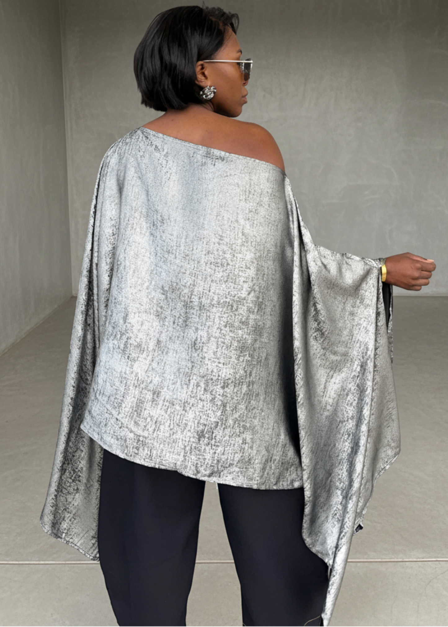 Person wearing a silver off-shoulder top and black pants in a minimalistic room.