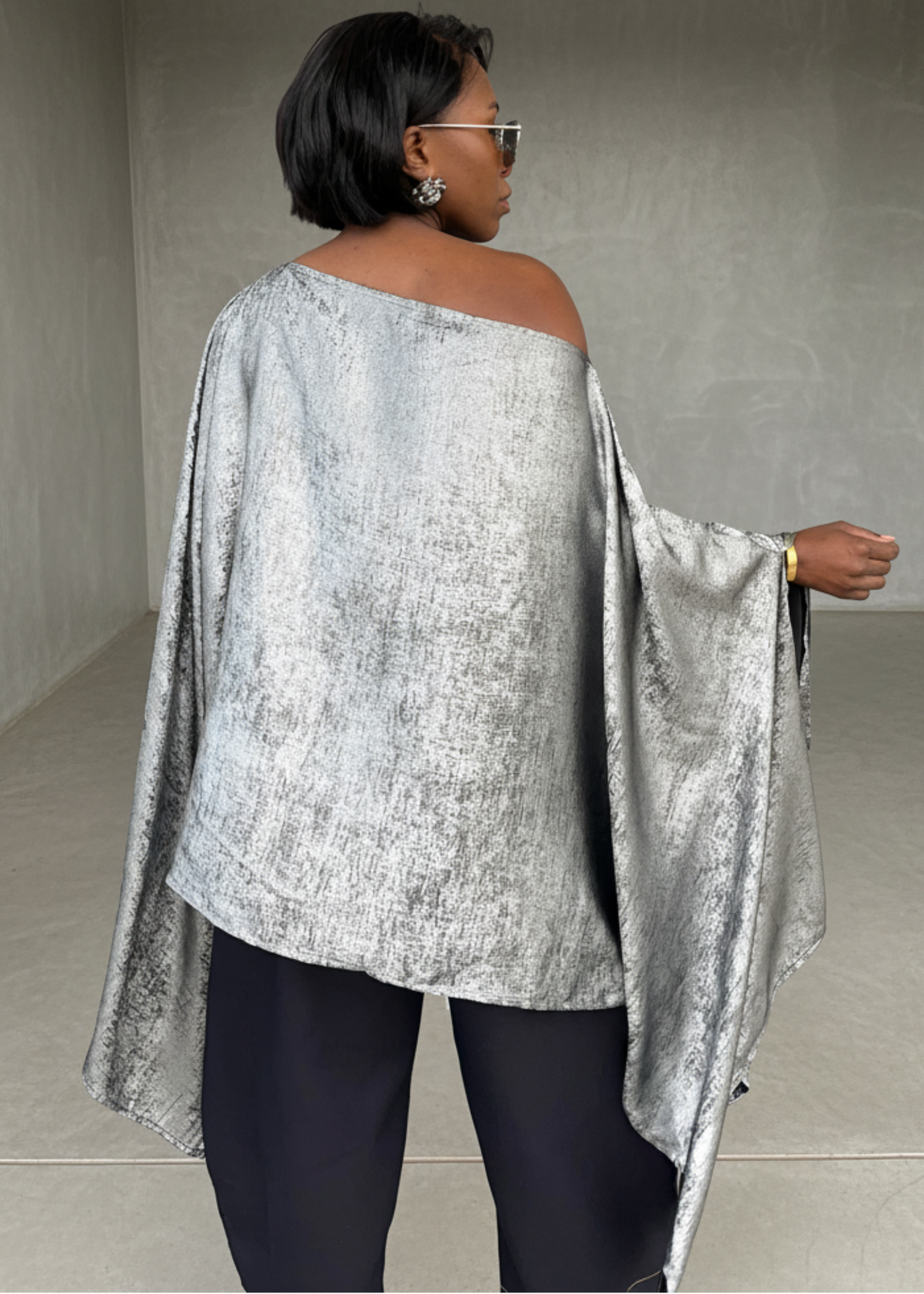 Person wearing a silver off-shoulder top and black pants in a minimalistic room.