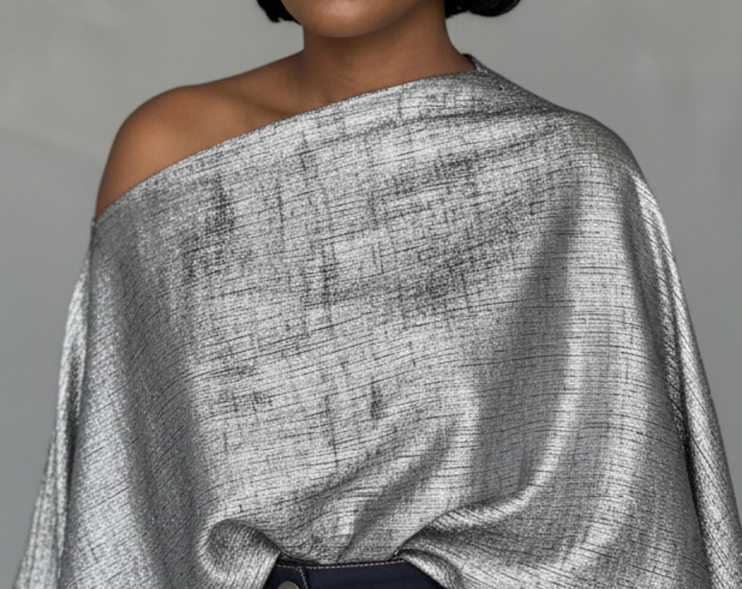 Woman wearing a silver off-shoulder top and dark pants against a gray background