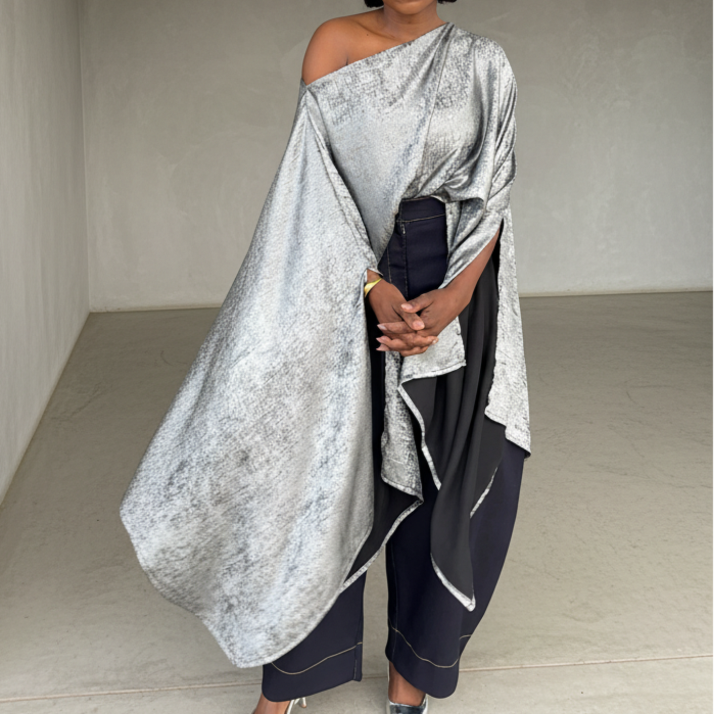Woman in a silver off-shoulder top and dark pants standing in a minimalistic room.