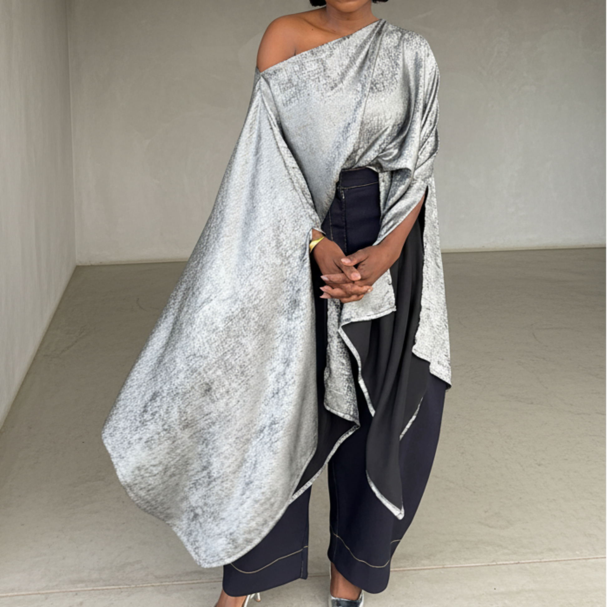 Woman in a silver off-shoulder top and dark pants standing in a minimalistic room.