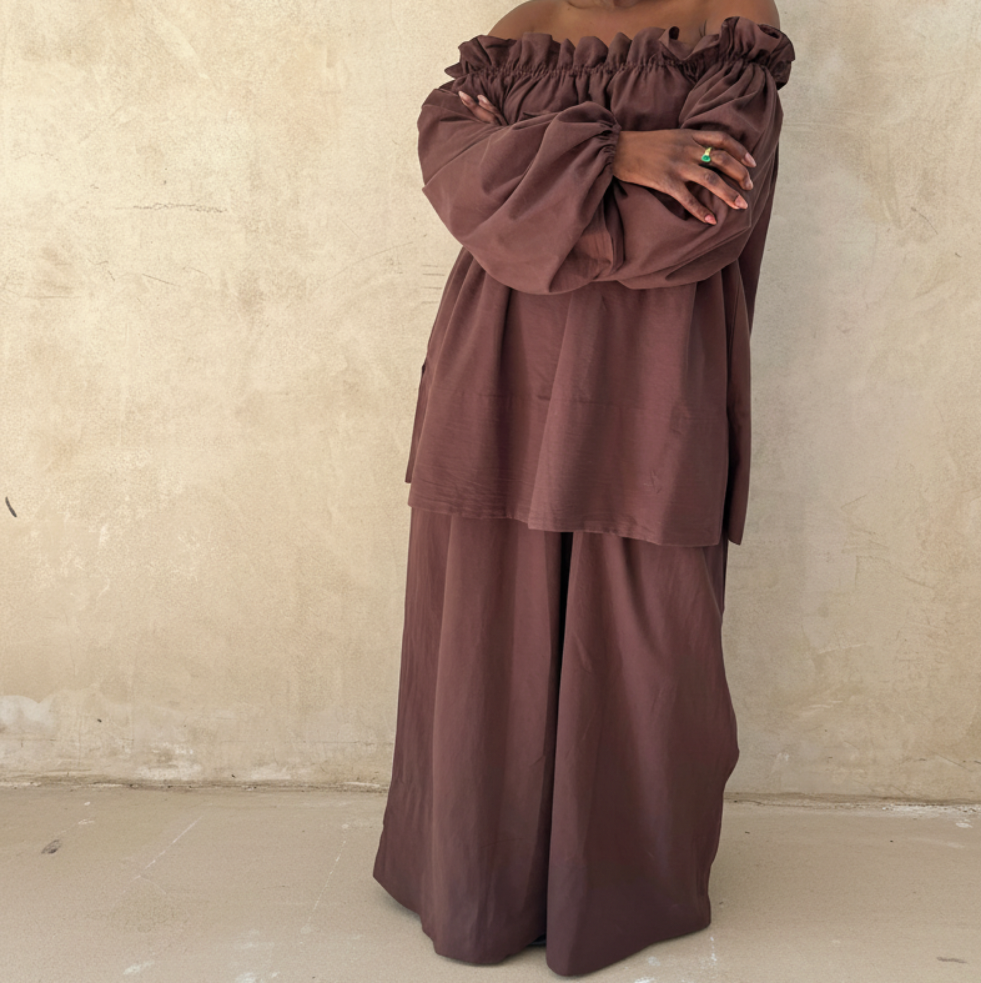Woman wearing a brown off-shoulder top and wide-leg pants against a beige wall.