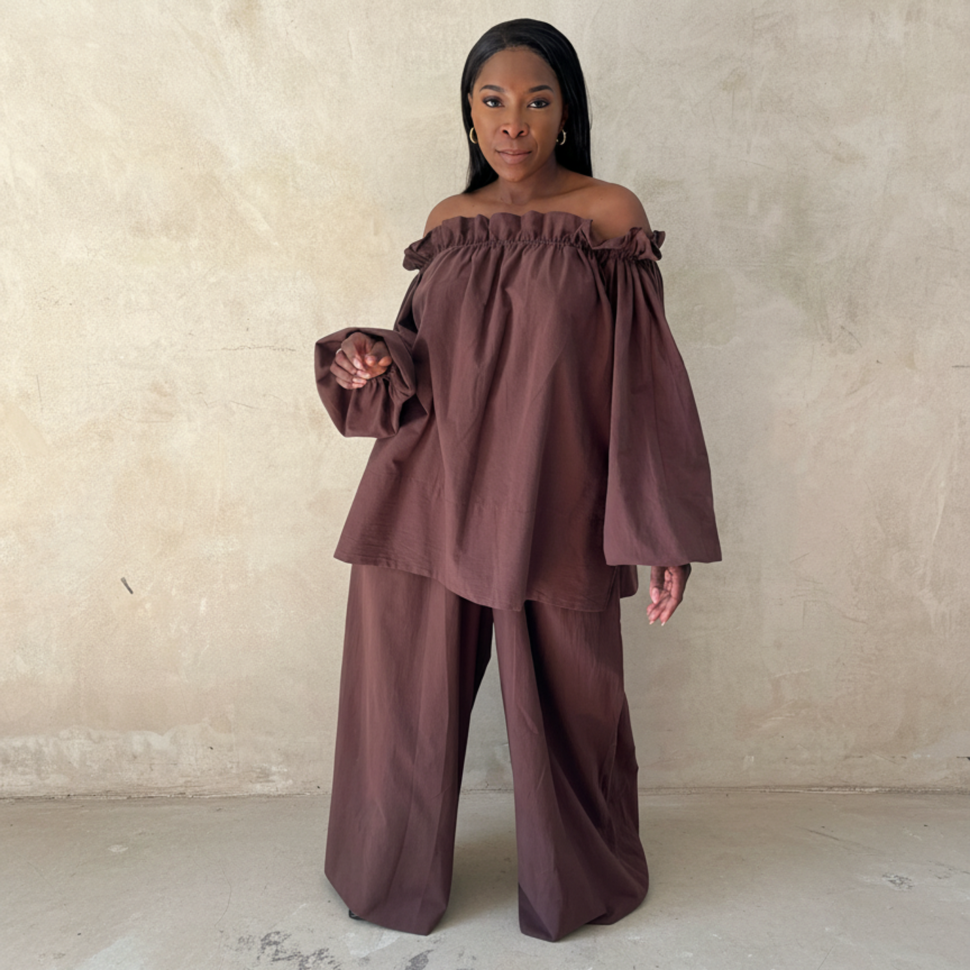 Woman wearing a brown off-shoulder top and wide-leg pants against a beige wall.